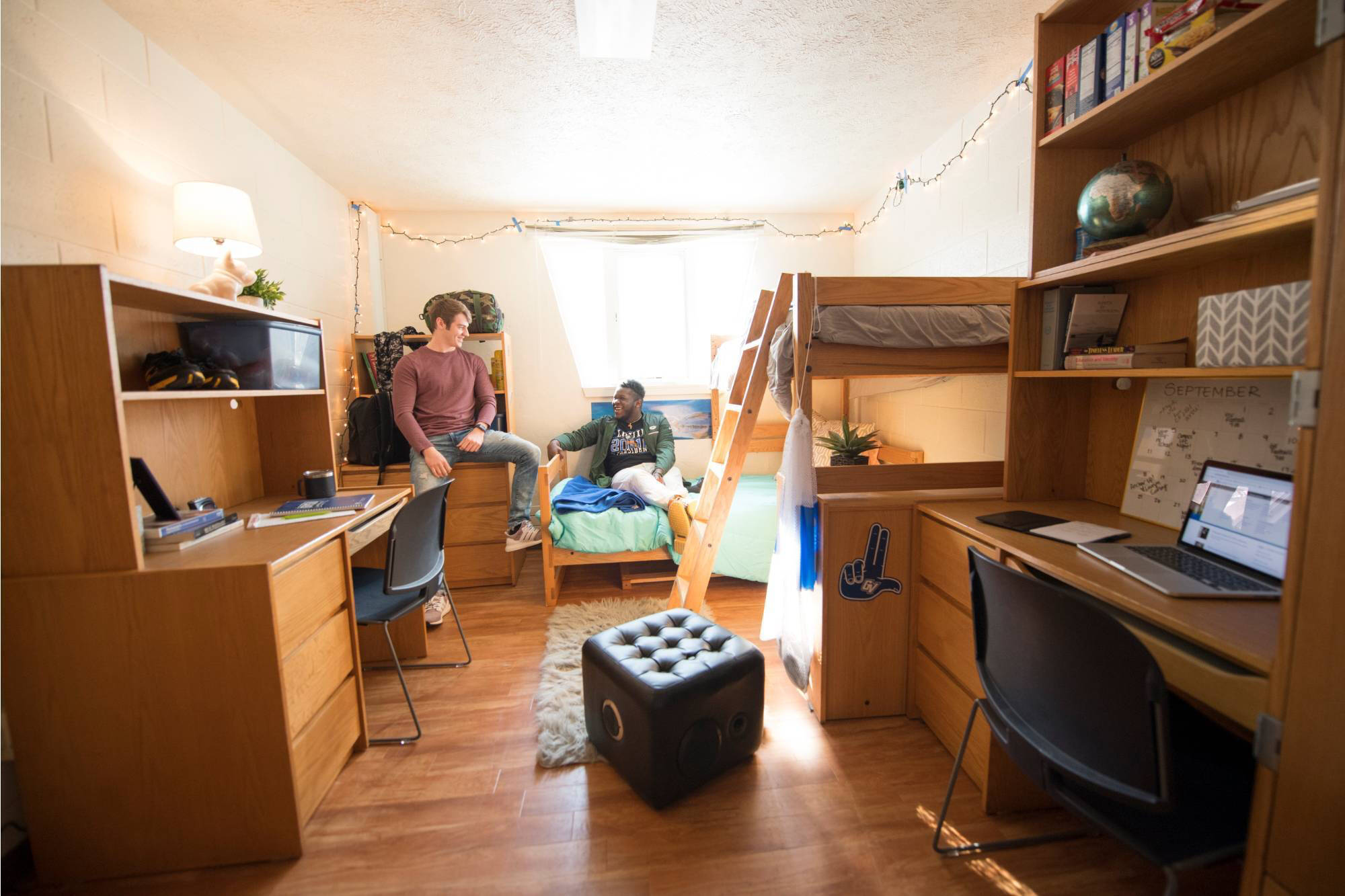 A bright Kistler room with wooden floors and bunk beds. Two students chat comfortably, surrounded by desks, laptops, books, and string lights. There's a cozy and inviting atmosphere.